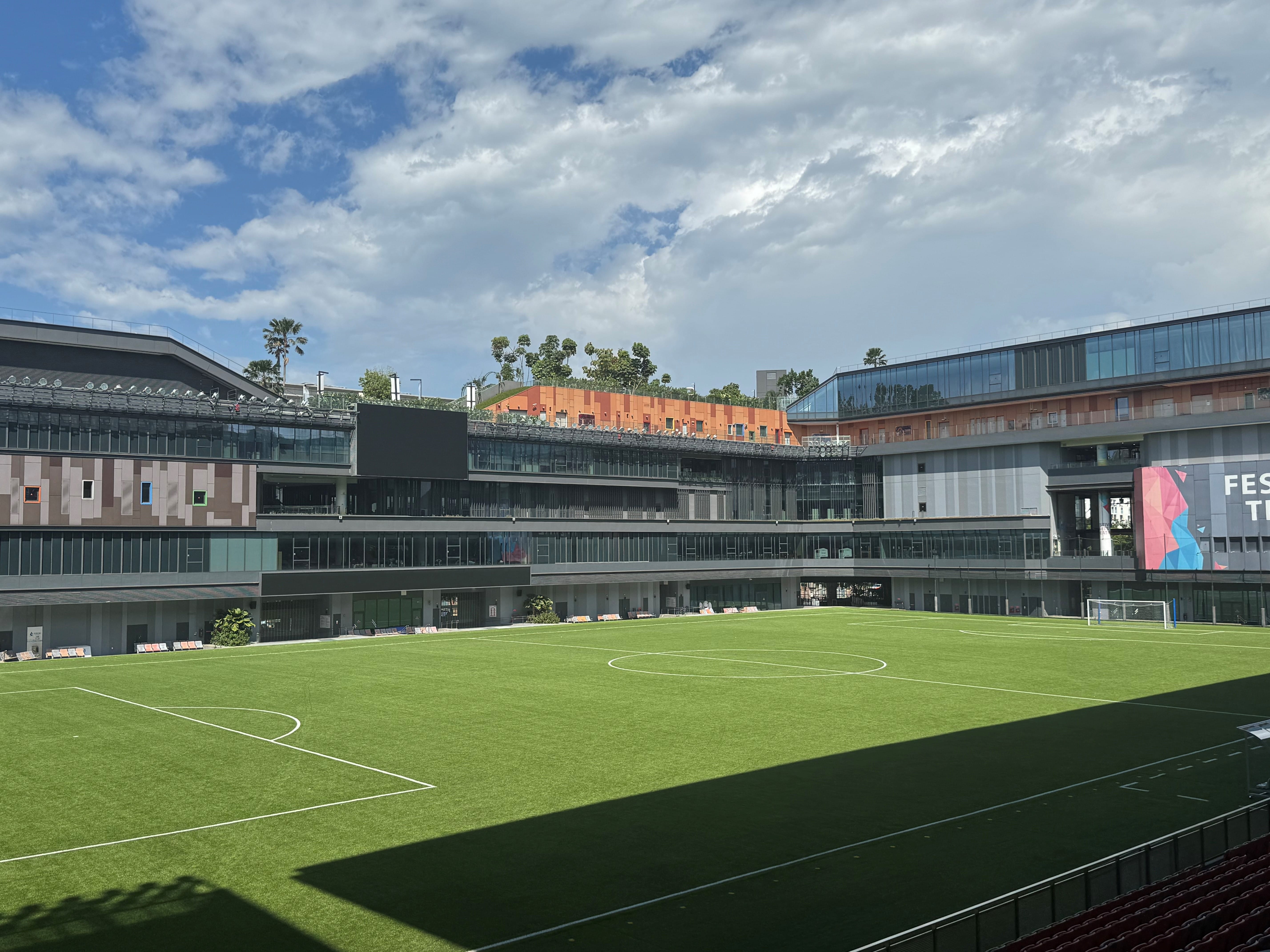 A modern school building surrounds a green soccer field under a blue sky with clouds.